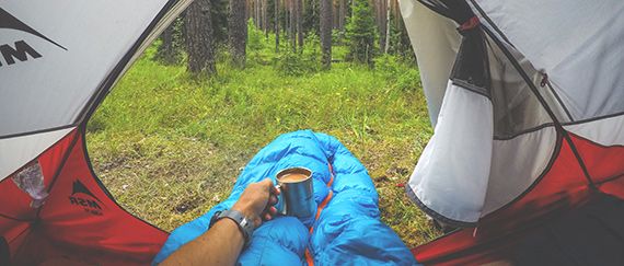 A man drinking coffee from a mug in a rented sleeping bag inside a hiking tent